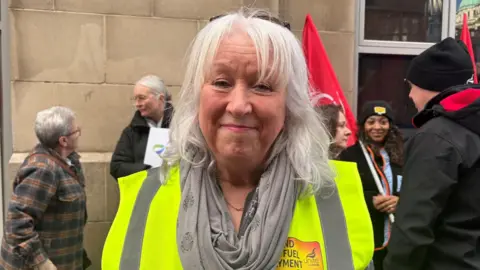 Dympna McGlade - a woman with shoulder-length grey hair is smiling at the camera as campaigners hold flags and talk in the background. She is wearing a yellow hi-vis jacket and a grey scarf.