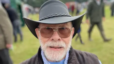 Rachel Davies Howard Ryder is looking at the camera in a close-up shot. He is wearing a black leather brimmed hat and sunglasses. He has a white beard and moustache