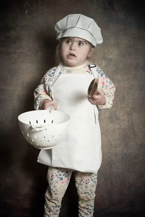 Debbie Todd Lacy-Mai, a young girl with brown hair, is wearing a white chef's hat and white apron over her floral jumper and trousers. She is holding a large wooden spoon in one hand and a white colander in the other.