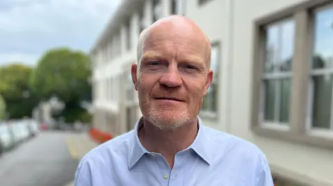 Deputy Gavin St Pier - A man with a bald head looking at the camera. He has a ginger and grey beard, and is wearing a blue button-up shirt with the collar open. 