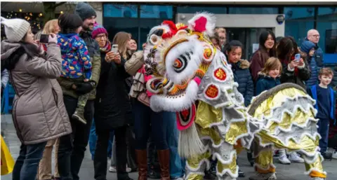 A multi-colured chinese dragong puppet is in front of a group of people of mixed ages standing next to eachother and smiling. 
