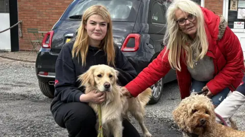 Three women kneel outside an industrial unit while holding three dogs. The unit has a large red sign on the front that reads "Beloved Doggie Holidays and Day Care". 