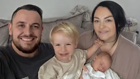 Man with short dark hair and beard with a dark top with a child with blonde hair and a beige top, a woman with brown hair and a brown top and a baby wrapped in white. They are sitting on a sofa.