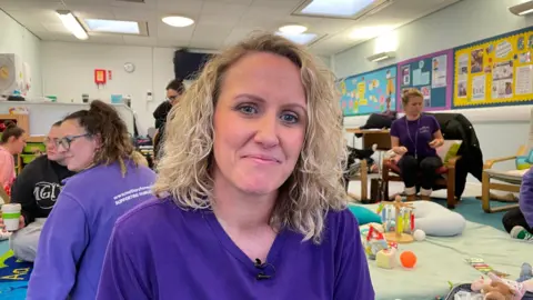 A woman with shoulder length, blond curly hair looks at the camera. She is wearing a purple t-shirt. Other women and a room full of children's toys is visible in the background.