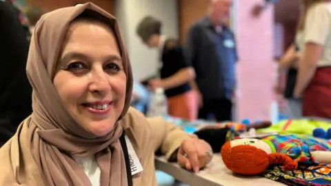 Amena Sadat smiles at the camera while bending down next to a table with a handmade doll