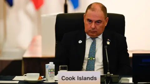 Getty Images Mark Brown sits in a chair looking serious. In front of him on a table are some cups, a microphone, and a tag that reads "Cook Islands".