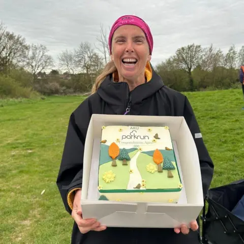 Apryl Hammett Apryl wearing a black coat and pink headband is pictured grinning and posing with a cake in a box. It is covered is icing showing a path through a field with trees, birds and the "parkrun" logo.