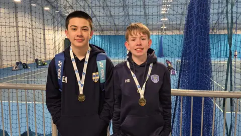 Two young boys in blue hooded tops with medals around their necks. There are indoor tennis courts behind them. 
