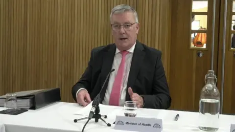 Mike Nesbitt at Stormont's Health Committee. He is sitting at a white table in front of a mic with a glass and bottle of water and a placing saying 'Minister of Health'. He is wearing glasses, a black blazer, white shirt and pink tie.