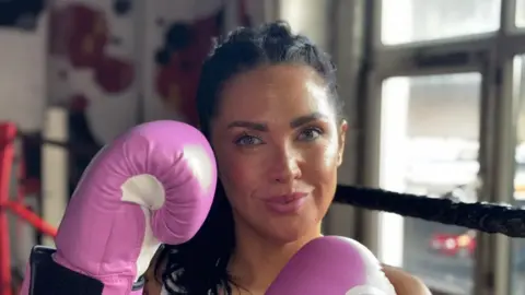 Woman with black hair looking at camera wearing pink boxing gloves 