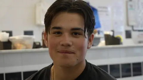 Jamie Niblock/BBC A young man wearing a black t-shirt and gold chain is sat in a cafe with the serving counter behind him