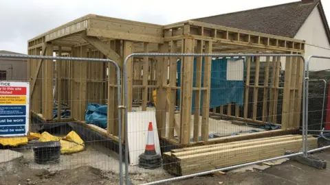 Marhamchurch Village Shop The one-story high timber frame of a building, during construction work. A temporary metal fence, building site signs, some traffic cones in the foreground.