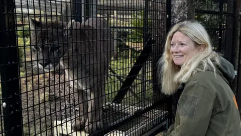 Wildside Exotic Rescue A woman with blonde hair is wearing a dark green coat and is sitting in front of an animal enclosure fence that is black. Behind it is a large lynx cat that is a brownish grey