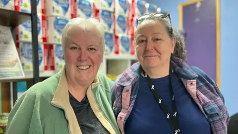 On the left is Yvonne Smith, she stands next to Sioux Watkins, who is the founder of Plate of Plenty. Smith has short white hair, wears a green fleece and a grey T-shirt. Watkins has grey hair tied in a pony tail with glasses on her head. She is wearing a tartan shirt, with a navy T-shirt underneath. Behind them is a row of toilet rolls.