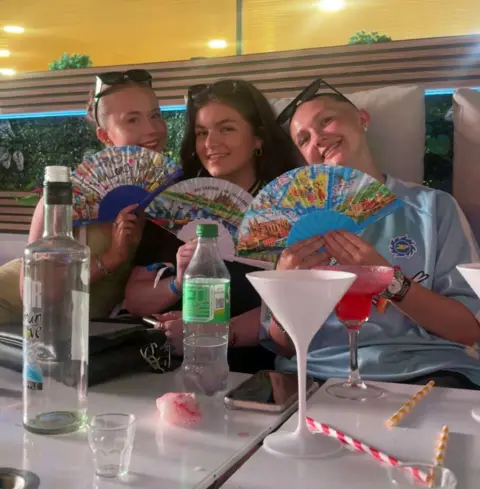 Taylor Coulter Three young women sitting in a bar, holding paper fans