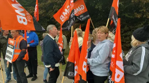 A dozen workers stand on a street holding orange and black "GMB" branded flags and signs. One of the signs reads: "OFFICIAL DISPUTE".