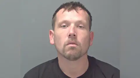 A head and shoulders mugshot image of Simon Loveridge. He is wearing a black T-shirt and standing in front of a grey background. He is looking gormlessly into the camera. 