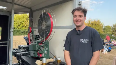 Festival organiser, Joe Cornick stands by the horse box with a film projector inside it - people can be seen setting up their chairs on the grass in the background