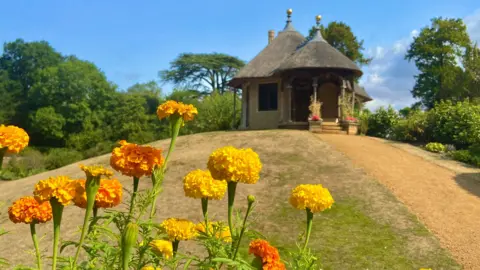 BBCWeatherWatchers/sunflowerdeb16 Yellow and orange marigold flowers on green stalks frame the picture of a thatched round cottage on top of a hill of dry grass and blue skies behind.