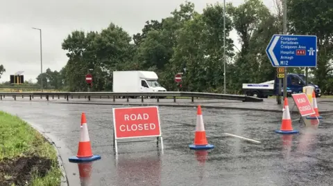 Red road closed sign beside traffic cones on the M12, a blue road sign showing the destinations of Craigavon and Portadown can be seen on the far right. A white van is in the distance.