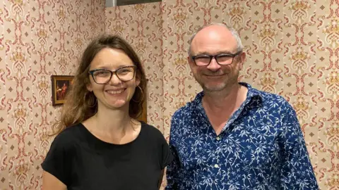 A woman with long hair and glasses standing next to a man with glasses who is wearing a blue shirt with a white flower pattern on it.