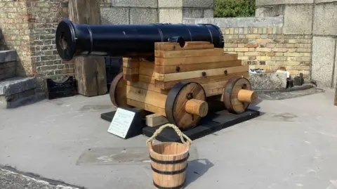 Alan Paulus A canon and wooden carriage. In the foreground is a small homemade bucket.