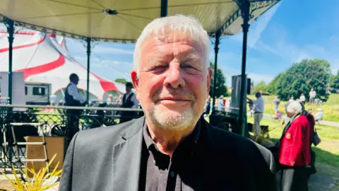 Claudia Sermbezis / BBC A man in a dark shirt and dark suit jacket looks at the camera smiling. Behind him there is a bandstand and people on the grass in the sunshine.
