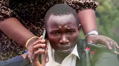 Duncan Wanga Felix Kemboi is comforted by a loved one who places her hands on his shoulders. He is visibly update as he holds a mobile phone to his ear. Image has been colour-corrected for distortion.