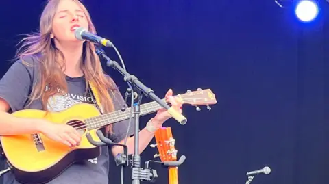 Nic Rigby/BBC Amelia Coburn plays a guitar and is singing on stage. She wears a dark-coloured T-shirt.