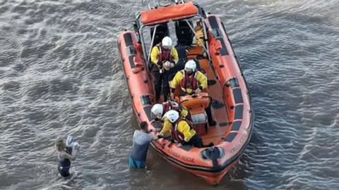 The orange Hornsea Rescue lifeboat alongside the two people in the water two crew members is bright yellow rescue gear and white helmets are helping a man into the boat with the woman waiting a few yards behind