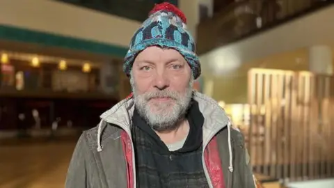 Kris Holland/BBC David Schaal in a brown duffel coat and a ragged blue bobble hat. 