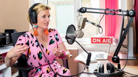 SuperFunkyPigeon Faye Dicker is sat at a desk with radio equipment surrounding her. She has a microphone on a stand and headphones on her desk. Behind her is a laptop an a red sign which reads "ON AIR". Faye has short blonde hair, wearing headphones and big orange earrings. Her dress is pink with multicoloured leopard spots 
