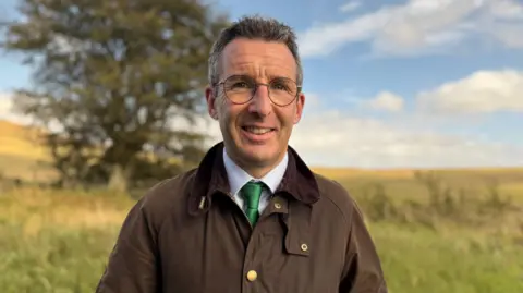 Andrew Muir standing in a field. He is wearing a brown coat, green tie and blue shirt. He has glasses on and has short black hair. There are trees and branches behind him. 