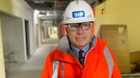 Image of Julian Auckland-Lewis. He is wearing a hi-vis jacket and hard hat. Ongoing construction can be seen behind him