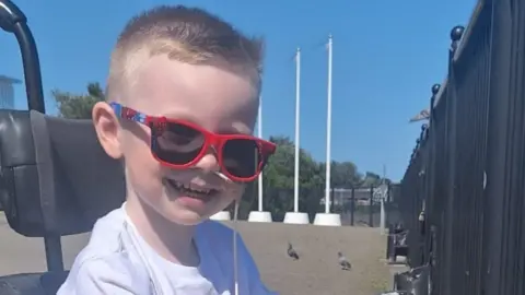 Dáibhin McConnell-Mailey, a four-year-old boy with short, blond hair, smiles at the camera while sitting in a wheelchair. He has red sunglasses on.