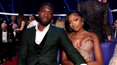 Getty Images Josh Oyinsan and Mimi Ngulube during the NTAs. Josh wears a achromatic  tuxedo with waistcoat and unbuttoned shirt, his manus  connected  Mimi's knee. Mimi wears an embellished nude dress. They're pictured astatine  the lawsuit   with a assemblage  seated down  them. 