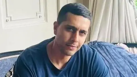 Kamran Aman wearing a blue top and sitting a sofa with a blue blanket behind him. He is looking at the camera and smiling. 