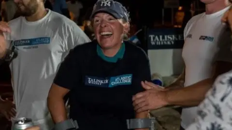 Atlantic Challenge/Ted Martin Kelda Wood in a baseball cap and black T-shirt, celebrating completing a crossing of the Atlantic with men wearing white T-shirts