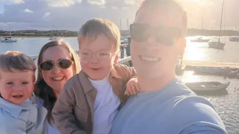 Nathan Davies A family of four, a man and woman wearing sunglasses and two small boys stand in front of a marina with boats in, taking a selfie.