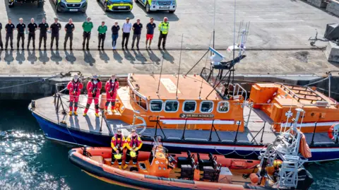 A group of emergency service personnel stand in a line near the edge of a dock. There are two boats pulled up to the dock, the one closer to the wall is bigger. Three people stand in orange jumpsuits on the deck of the larger vessel. Two people in yellow and black overalls stand on the deck of the smaller boat. They are all looking at the camera.