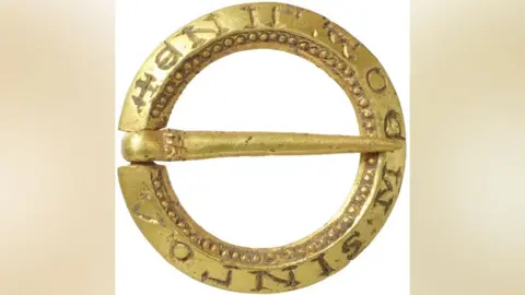 Andrew Williams/Norfolk County Council A close-up of a small, gold round medieval brooch. It is made from a ring with a gold pin crossing over its middle. The outer ring has words etched into it and the inner ring has a decoration of tiny gold dots