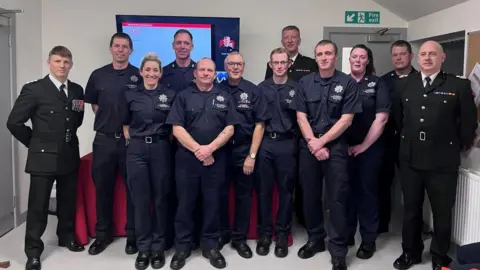 Office of Cumbria's Police, fire and crime commissioner A dozen people in fire service and staff uniforms stand to pose for a photo in the refurbished fire station. 