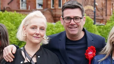 PA Media Andy Burnham, wearing glasses and a blue jacket and T-shirt with a red Labour rosette, and Marie-France Van Heel, with blonde hair and wearing a black top, smile at the camera