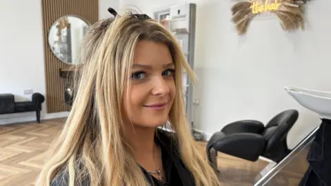 A woman with blonde hair sits in a salon chair looking at the camera.