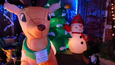 Santa's Garden An inflatable reindeer with a sign around its neck reading "Happy Xmas my name's Twinkle" stands next to an inflatable Christmas tree and an inflatable snowman.