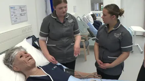 The image shows a clinical training ward where healthcare students in uniform are gathered around a hospital bed, practising care on a lifelike patient mannequin connected to monitoring equipment. The space is bright and modern, with another simulated patient visible in a bed behind them, reinforcing the realistic hospital setting.