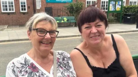 BBC/ Emily Johnson Two women sit on a bench on a high street in Acomb, York.