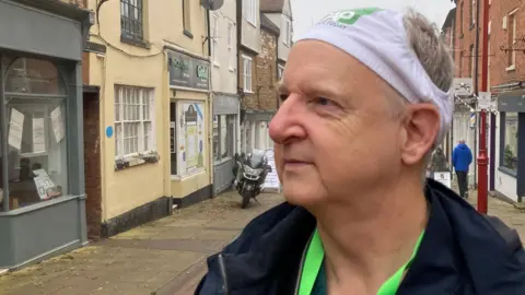 Mark Robson with short grey hair and a blue raincoat wearing white pants on his head with a green Christians Against Poverty logo. He is looking at shops to his right. There are shops on both sides of a pedestrian street and some shoppers are walking down it.