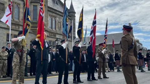 Military personnel stand in a line holding up flags