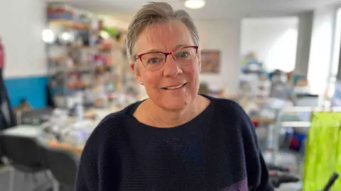 Mary Hervé pictured with short grey hair wearing a purple jumper. She has pearl earrings on and glasses with a red frame.